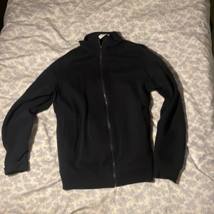 Svart zip-kofta från Cortefiel - Svart kofta från Cortefiel med hel dragkedja framtill och ståkrage. Tröjan har långärmade ärmar och en clean, stilren look utan mönster eller extra detaljer. Perfekt för dig som gillar enkel och klassisk stil.