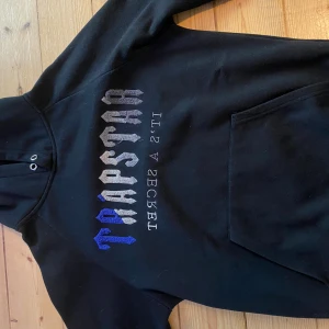 Svart hoodie från Trapstar med tryck - Svart hoodie från Trapstar med stor broderad logga i grått och blått på bröstet. Klassisk känguruficka framtill och huva med snörhål. Perfekt för dig som gillar streetwear och vill sticka ut med en fet logga.
