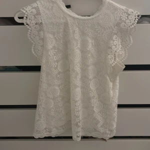 Vit piecesblus  - Supersöt vit blus från pieces med detaljerad spets och blommigt mönster. Blusen har v-ringning bak, korta ärmar med volangkant och snygga broderade detaljer. Perfekt för dig som gillar romantisk och feminin stil. Passar till både jeans och kjol!