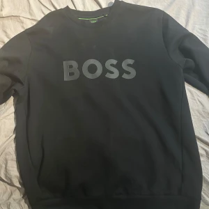 Svart BOSS sweatshirt i XL - Svart sweatshirt från BOSS i storlek XL med stor logga framtill. Tröjan har rund halsringning och långa ärmar. Perfekt för dig som gillar stilren och enkel design med en tydlig logga. Skön och mjuk känsla, passar till många outfits.