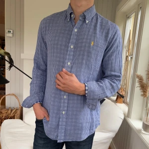 Blårutig skjorta från Polo Ralph Lauren - Säljer en klassisk blårutig skjorta från Polo Ralph Lauren i bomull. Skjortan har button-down krage, lång ärm och vit broderad logga på bröstet. Custom fit-modell som sitter snyggt och avslappnat. Perfekt till jeans eller chinos.