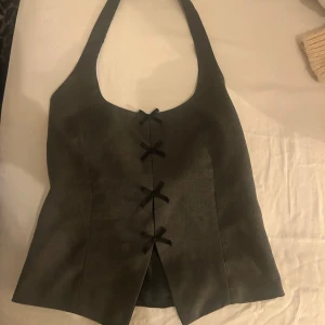 Grå omlottväst från Zara med snörning - Snygg grå väst från Zara i omlottmodell med breda axelband och dekorativa svarta snörningar framtill. Västen har en stilren, figurnära passform och är tillverkad i ett mjukt tyg som ger en modern vibe. Perfekt att styla över en skjorta eller t-shirt.