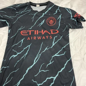 Manchester City Haaland #9 fotbollströja S - Snygg Manchester City fotbollströja med Haaland och nummer 9 på ryggen. Tröjan är svart med ljusblå blixtmönster och röda detaljer, inklusive klubbmärke och Etihad Airways-logga. Kortärmad och tillverkad i lätt, ventilerande polyester. Perfekt för dig som vill sticka ut på planen.