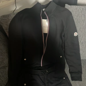 Svart Moncler bomberjacka med dragkedja - Svart bomberjacka från Moncler med klassisk logga på ärmen och snygg randig detalj längs dragkedjan. Jackan har ribbade muddar, knappar vid fickorna och sportig look. Perfekt för dig som vill ha en stilren och exklusiv jacka med streetkänsla.