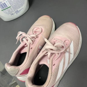 Adidas rosa sneakers med vita ränder - Snygga Adidas sneakers i ljusrosa med klassiska vita ränder på sidan. Skorna har mesh-överdel för bra ventilation, snörning och platt sula med Lightmotion-dämpning. Perfekta för träning eller chill häng. Färgen är ljusrosa med detaljer i vitt och mörkrosa. Sköna och lätta på foten.