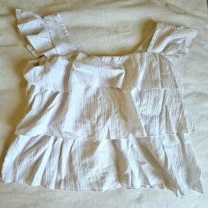 Vit volangblus med breda axelband - Säljer en söt vit blus med breda axelband och volangdetaljer. Toppen har en luftig och flowy passform med flera lager volanger som ger en romantisk vibe. Perfekt för varma dagar och att styla med jeans eller kjol.