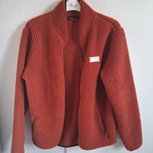 Rostbrun teddyjacka från True North - Mysig teddyjacka i rostbrunt från True North, storlek M. Jackan har lång ärm, hög krage och två fickor med dragkedja. Perfekt för kyliga dagar och har en riktigt skön, fluffig känsla. Snygg och enkel att matcha med jeans eller hoodies.