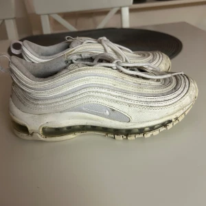 Nike Air Max 97 vita sneakers - Nike Air Max 97 sneakers i helvitt med ikoniska vågformade linjer och synlig Air-sula, storlek 37,5. Skorna har snörning och en platt sula, tillverkade i syntet och textil. 