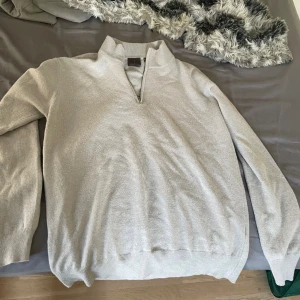 Ljusgrå half zip-tröja Oscar Jacobson - Snygg ljusgrå stickad tröja från Oscar Jacobson med half zip och hög krage. Tillverkad i Italien och har ribbade muddar vid ärmslut och nederkant. Perfekt för lager-på-lager och stilrena outfits.