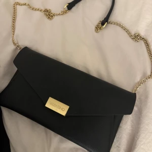 Svart kuvertväska från Valentino - Stilren svart kuvertväska från Valentino med elegant guldfärgad kedja och metallplatta med logga framtill. Väskan har en klassisk, rektangulär form och är tillverkad i skinn med slät finish. Perfekt för att lyfta din outfit med en lyxig touch.