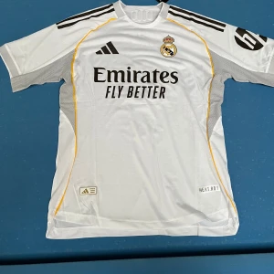 Real Madrid Mbappé fotbollströja  - Snygg vit Real Madrid fotbollströja från Adidas med Mbappé och nummer 9 på ryggen. Kortärmad modell med svarta detaljer och gula linjer längs sidorna. Officiell klubbmärke på bröstet och sponsortryck 'Emirates Fly Better'. Tillverkad i lätt och ventilerande material. Aldrig använd. Lapp finns kvar.