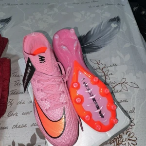 Nike Mercurial fotbollsskor - Säljer ett par Nike Mercurial fotbollsskor i en snygg rosa färg med orange och svart swoosh. Skorna har snörning, platt sula med dobbar och är tillverkade i syntetmaterial. Perfekta för dig som vill sticka ut på planen!