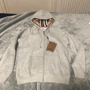 Grå hoodie från Burberry med dragkedja - Snygg ljusgrå hoodie från Burberry med klassiskt rutigt foder i huvan. Tröjan har dragkedja framtill, snörning vid huvan och två fickor. Perfekt för dig som vill ha en stilren och exklusiv look med en touch av Burberrys ikoniska design.