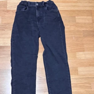 Svarta jeansbyxor från Kappahl, stl 170 - Svarta jeansbyxor från Kappahl i storlek 170. Modellen har hög midja med resår baktill, klassiska fem fickor och raka ben. Snygga kontrastsömmar och knapp med denim-detalj framtill. Perfekta för en avslappnad och trendig look.