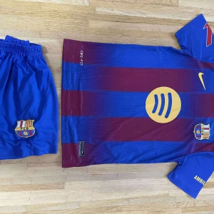 FC Barcelona Raphinha matchtröja Nike - Säljer en officiell FC Barcelona fotbollströja med Raphinha 11-tryck. Tröjan är kortärmad, har breda blå och vinröda ränder, gula detaljer och klubbmärke på bröstet. Materialet är lätt och andas, perfekt för match eller träning. Shorts medföljer, även de med klubbmärke.
