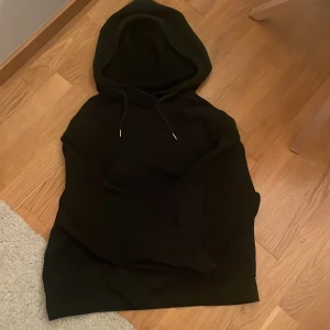Svart hoodie från Lindex med huva - En svart hoodie från Lindex med klassisk huva och snörning. Tröjan har lång ärm och är tillverkad i mjukt material, perfekt för chill dagar. Enkel och stilren design utan tryck eller mönster. Andvänd ett fåtal gånger men färgen har gått bort lite i tvätten 