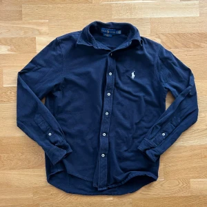 Mörkblå skjorta från Ralph Lauren - Snygg mörkblå skjorta från Ralph Lauren med klassisk krage och vita knappar. Materialet är lätt och luftigt mesh i bomull, perfekt för dig som gillar stilrena plagg med diskret broderad logga på bröstet. Långärmad modell med normal passform.