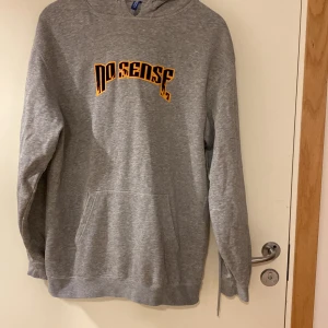 Grå hoodie med flames-tryck No Sense - Grå hoodie från Gildan med huva och känguruficka. På bröstet står det 'No Sense' i svart text med orange flames-effekt. Skön passform och perfekt för chill dagar. Klassisk streetstyle-vibe.
