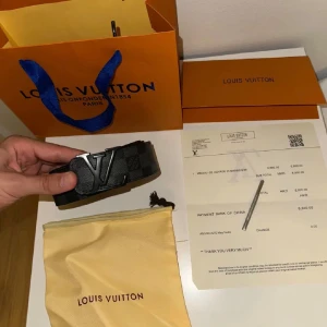 Louis Vuitton svart bälte - Snyggt svart bälte från Louis Vuitton med klassiskt rutigt mönster och stor LV-logga i metall på spännet. Bältet är gjort i exklusivt skinn och levereras med originalpåse och kvitto. Perfekt accessoar för att lyfta din outfit.