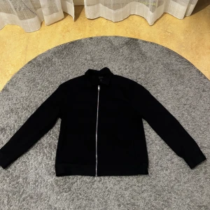 Svart overshirt med dragkedja - Stilren svart overshirt med hel dragkedja framtill och klassisk krage. Jackan har raka ärmar och en minimalistisk look som passar perfekt till streetwear eller en clean outfit. Materialet känns mjukt och lätt, perfekt för lager-på-lager under vår och höst.