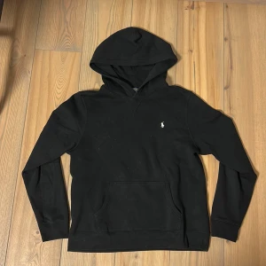 Svart hoodie från Polo Ralph Lauren - Svart hoodie från Polo Ralph Lauren med klassisk huva och känguruficka. Tröjan har en liten vit broderad logga på bröstet och är tillverkad i mjuk bomull. Perfekt för en chill och stilren look.