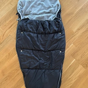 Nomad åkpåse - Varm och mjuk åkpåse från Nomad, svart utsida och grå fleece på insidan. Passar barnvagn, med flera öppningar för sele. Dragkedja och tryckknappar för enkel användning. Fint  skick, inga synliga defekter eller fläckar.