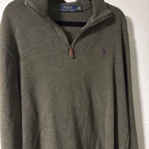 Olivgrön half zip tröja Polo Ralph Lauren - Olivgrön långärmad tröja från Polo Ralph Lauren med lila broderad logga på bröstet. Tröjan har en snygg half zip-dragkedja med läderdetalj och stårkrage. Tillverkad i mjuk bomull, perfekt för lager-på-lager under höst och vinter.