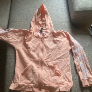 Ljusrosa Adidas hoodie med dragkedja - Snygg ljusrosa hoodie från Adidas med klassiska vita ränder längs ärmarna och logga på bröstet. Tröjan har huva, fickor framtill och den har mjukt bomullsmaterial och långärmad modell.