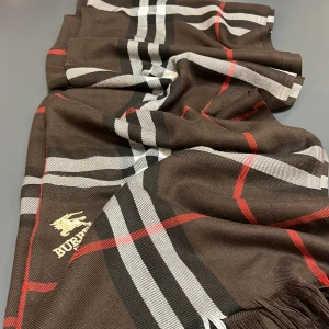 Brun rutig halsduk från Burberry - Snygg halsduk från Burberry med klassiskt rutigt mönster i brunt, svart, vitt och röda linjer. Halsduken har fransar i ändarna och ett broderat Burberry London-emblem i guld. Perfekt accessoar för att lyfta din outfit med lyxig känsla.