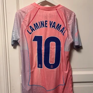 FC Barcelona Lamine Yamal Nike tröja - Säljer en FC Barcelona fotbollströja från Nike med Lamine Yamal och nummer 10 på ryggen. Tröjan är kortärmad och har en mix av rosa och ljusblå färger med tunna vita linjer. Materialet är Dri-FIT ADV för bästa komfort. FCB-märke och Spotify-logga på bröstet.