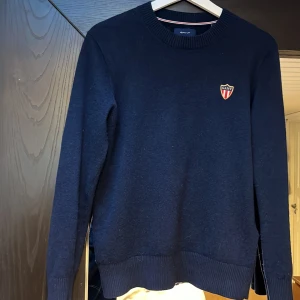 Mörkblå stickad tröja från Gant - Snygg mörkblå stickad tröja från Gant med rund halsringning och broderad Gant-logga på bröstet. Tröjan har ribbade muddar vid ärmslut och nederkant, perfekt för dig som gillar klassisk och stilren look.