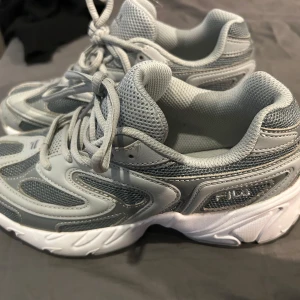 Grå sneakers från Fila - Säljer ett par snygga Fila sneakers i ljusgrå och vit färg med mesh och syntetpaneler. Skorna har rund tå, snörning och en chunky, vit platt sula. Perfekta för dig som gillar sportig stil och vill ha bekväma skor med cool design.
