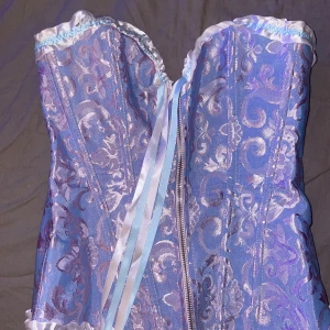 Ljuslila korsett med satinrosett - Superfin ljuslila korsett med glansigt, brokadliknande mönster och vita samt ljusblå satinband framtill. Toppen har söt volangkant upptill och nedtill samt dragkedja framtill. Perfekt för dig som vill sticka ut med en romantisk och trendig look.