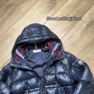 Mörkblå pufferjacka från Aumile - Snygg mörkblå pufferjacka från Aumile med huva och dragkedja. Jackan har ett rutigt innerfoder i blått, rött och vitt som ger en schysst kontrast. Perfekt för kalla dagar när du vill hålla stilen och värmen. Slut såld i webben oanvänd 