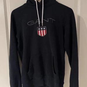 Svart hoodie från Gant med brodyr - Svart hoodie från Gant med vit snörning i huvan och broderad logga samt sköld med stjärnor och ränder på bröstet. Klassisk känguruficka framtill och långa ärmar. Perfekt för en chill och sportig look.