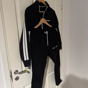 Palm Angels set - Svarta mjukisbyxor från Palm Angels med vita sidoränder och snörning i midjan. Byxorna har en slim passform och är tillverkade i mjukt material med logotypen tryckt på framsidan. Perfekt för en chill och sportig look. Palm angels tröja med vita ränder. Kom med frågor pris ej fast