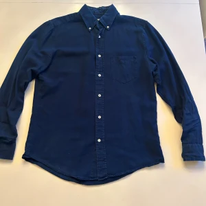 Mörkblå linneskjorta från GANT i storleken M - Snygg mörkblå linneskjorta från GANT Rugger i storlek M. Klassisk modell med button-down krage, bröstficka och vita knappar framtill. Skjortan är långärmad och tillverkad i mjuk bomull, perfekt för en clean och chill stil.