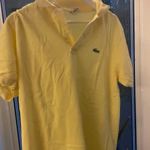 Gul pikétröja från Lacoste - Snygg gul pikétröja från Lacoste med klassisk krage och två knappar framtill. Tröjan har korta ärmar och den ikoniska gröna krokodilloggan på bröstet. Perfekt för dig som gillar stilrena och sportiga plagg.