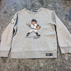 Grå sweatshirt Polo Bear Ralph Lauren - Grå sweatshirt från Polo Ralph Lauren med ikoniska Polo Bear i New York Yankees-baseballoutfit på bröstet. Stort Yankees-märke på ärmen och patch nertill. Klassisk rund hals och långa ärmar.  Storlek M (10-12 år)