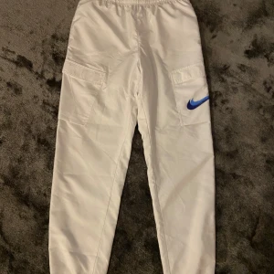 Vita cargopants från Nike - Säljer ett par vita cargopants från Nike med blå swoosh-detalj på benet. Byxorna har elastisk midja och stora fickor på sidorna. De är tillverkade i ett lätt bomullsmaterial och har en avslappnad passform som är perfekt för en sportig look.