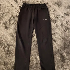 Svarta mjukisbyxor från Champion - Svarta mjukisbyxor från Champion med vit logga på vänster ben. Byxorna har elastisk midja med vit snörning och två sidofickor. Tillverkade i mjukt material som är perfekt för chill eller träning. Klassisk och enkel design som funkar till det mesta.