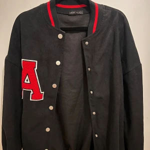 Svart bomberjacka med röd detalj SHEIN - Svart bomberjacka från SHEIN med stora röda bokstaven A på bröstet. Jackan har tryckknappar framtill, ribbade muddar med röda ränder och klassisk college-stil. Perfekt för dig som gillar sportig och trendig look.