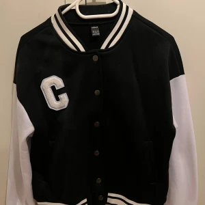 Svartvit bomberjacka från SHEIN - Snygg svartvit bomberjacka från SHEIN med klassisk college-look. Jackan har vita ärmar, svart kropp, randiga muddar och en stor broderad C på bröstet. Stängs med knappar framtill och har två fickor. Perfekt för en sportig och trendig stil.