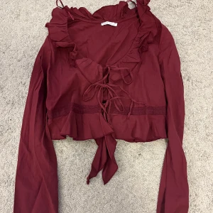 Vinröd blus med volang från Bershka - Supersöt vinröd blus från Bershka med långa ärmar, volangkant runt ringningen och knytning framtill. Croppad modell med romantiska detaljer och lätt material som känns luftigt. 