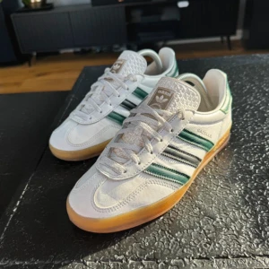 Adidas Gazelle vita och gröna sneakers - Säljer ett par klassiska Adidas Gazelle sneakers i vitt skinn med gröna och svarta ränder på sidorna. Skorna har gummisula i brunt, snörning och rund tå. Ikonisk retrostil med Adidas-logga på plösen och hälen. Skriv om ni har funderingar, använda ungefär 5 gånger bara!