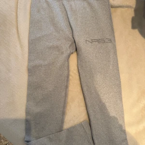 Grå ribbade leggings från New Apparel - Snygga grå tights från New Apparel i ribbad, stretchig polyesterblandning. De har en tight passform och hög midja, med diskret logga på benet. Perfekta för chill eller träning, och superbekväma tack vare det mjuka materialet.