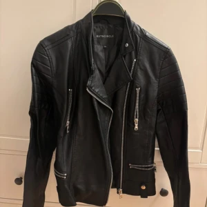 Svart skinnjacka från Rut&Circle XS - Snygg svart skinnjacka från Rut&Circle i storlek XS. Jackan har coola detaljer med flera dragkedjor, quiltade partier på axlarna och klassisk bikerkrage. Perfekt för dig som vill ha en edgy look med stilrena vibbar.