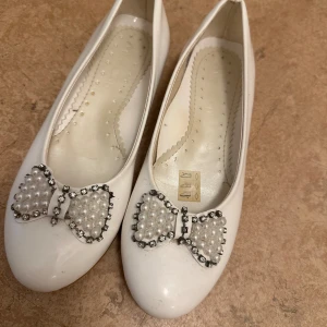 Vita ballerinaskor med pärlrosett  - Snygga vita ballerinaskor med rund tå och platt sula. Framtill sitter en dekorativ rosett täckt av pärlor och glittrande stenar som ger en extra touch. Skorna har en klassisk och feminin look, perfekta för att lyfta din outfit.