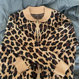 Leopardmönstrad stickad kofta - Säljer en snygg stickad kofta med leopardmönster i beige, svart och brunt. Koftan har dragkedja framtill, ribbade muddar och krage. Perfekt för dig som vill sticka ut lite extra med din outfit.