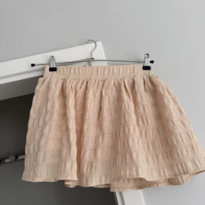Beige kortkjol från Zara - Supersöt beige kortkjol från Zara med voluminös form och elastisk midja. Kjolen har ett diskret rutigt mönster och är gjord i ett mjukt bomullstyg som känns luftigt och bekvämt. 